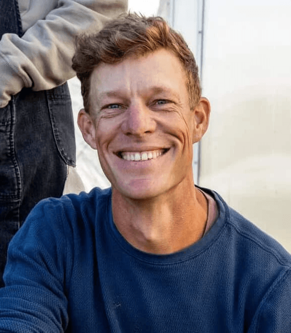 A woman with shoulder-length brown hair, wearing a white shirt, smiles against a blurred outdoor background.