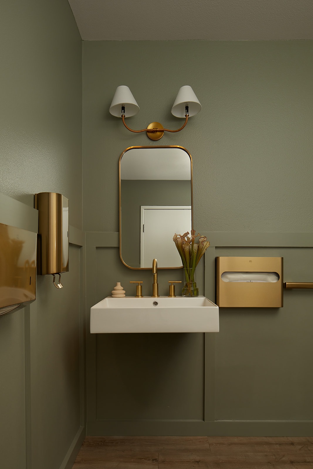Modern washroom interior at DC Dermatology featuring gold fixtures, wall-mounted sink, brass mirror, and soft green walls designed to feel calm and elevated.