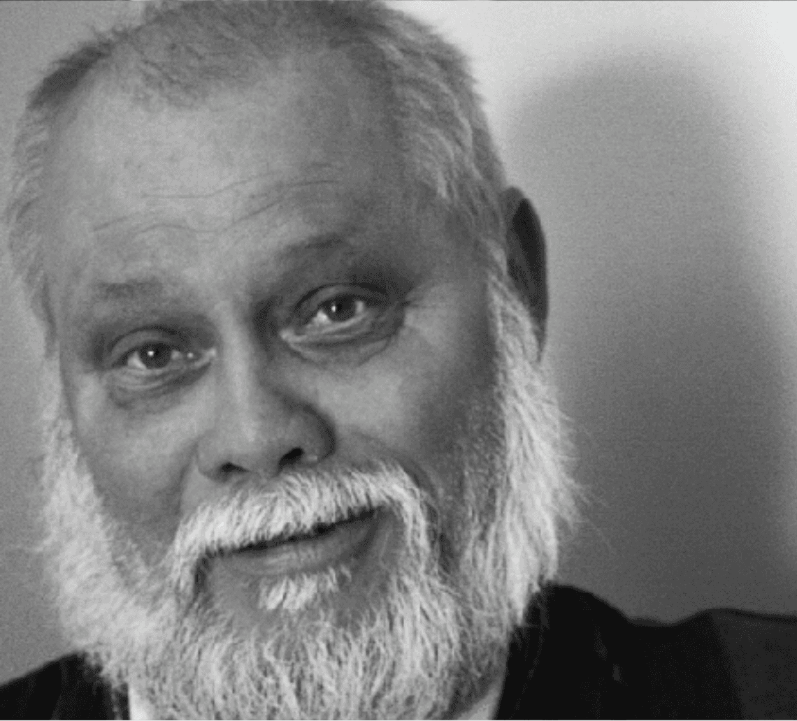 A smiling older man with a beard, wearing a dark shirt, against a light background. The image is in black and white.