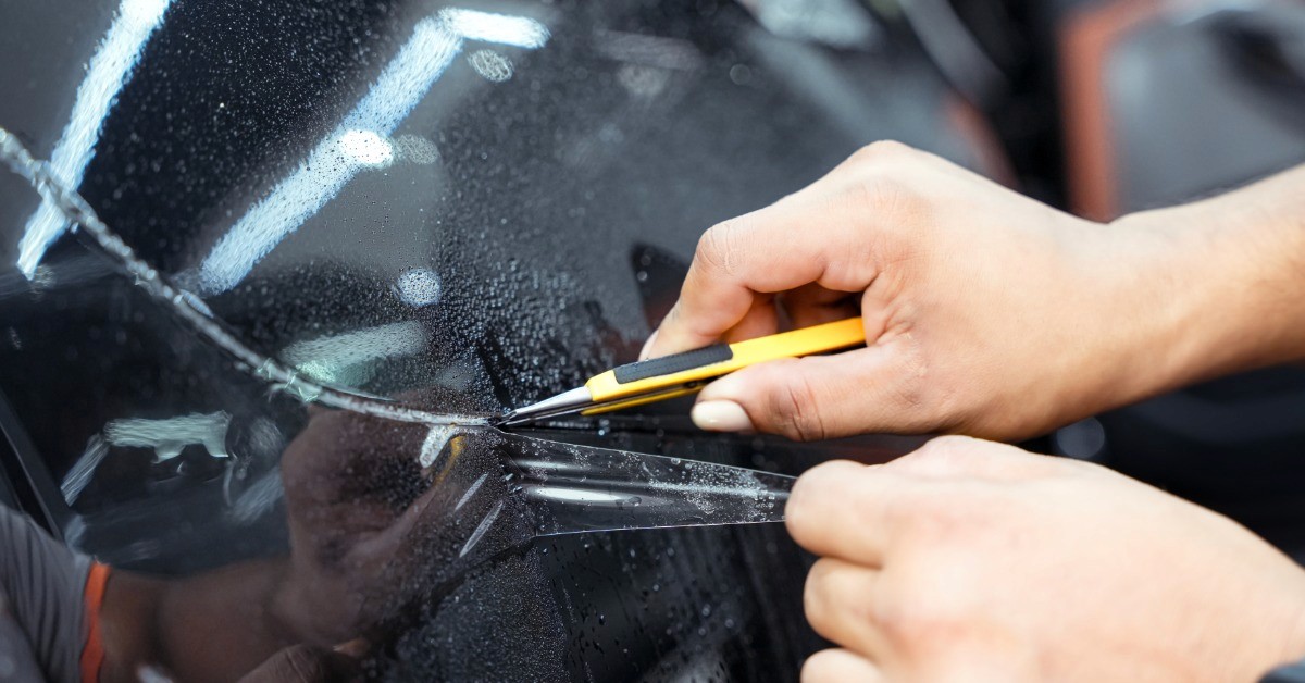Hands trimming clear protective film on a black car panel with a yellow utility knife and water droplets nearby.