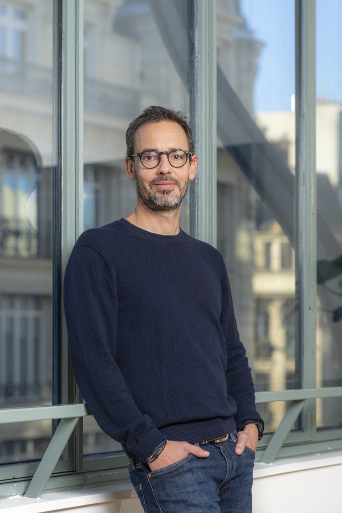 Young person with short hair wearing a dark flat cap, black cardigan, and white turtleneck, exuding a calm and composed expression against a plain background.