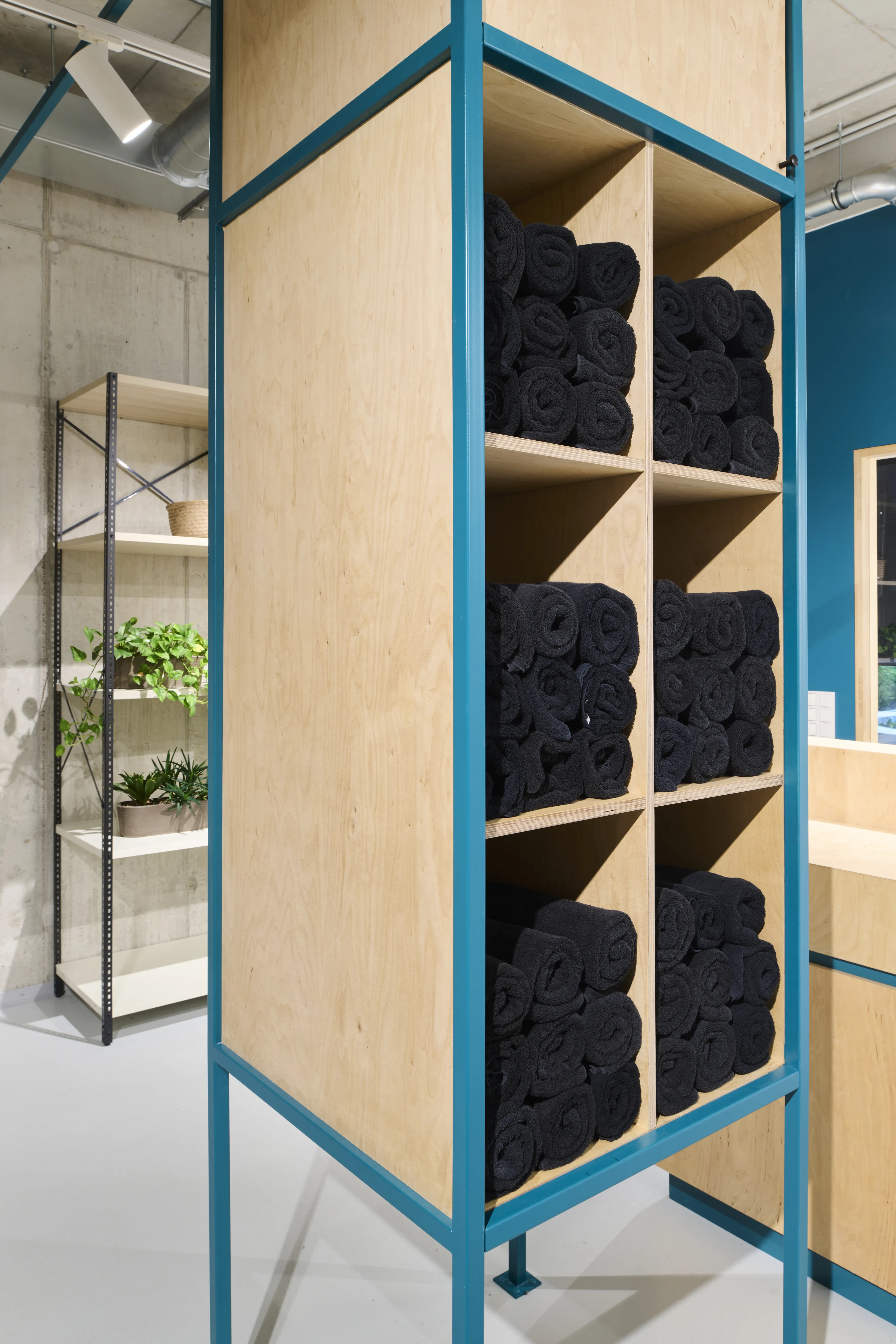 Tall plywood shelf in a teal steel frame at HELLOFRESH GYM filled with rolled black towels, next to a plant shelf against raw concrete.