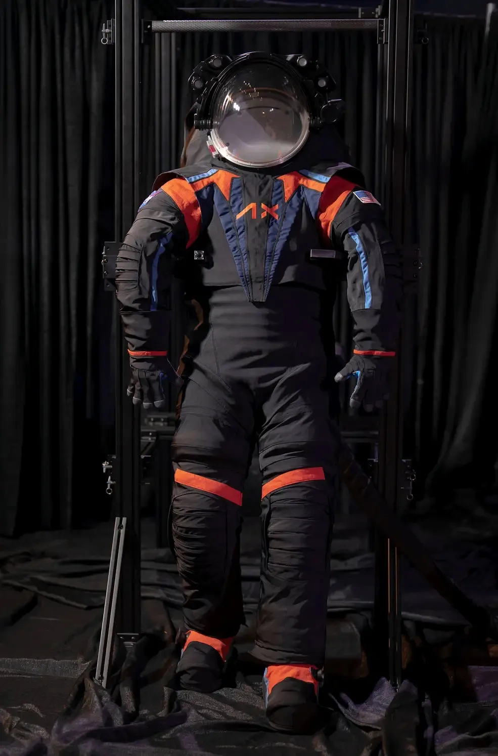 A futuristic astronaut suit, featuring black material with vibrant orange and blue accents, is displayed against a dark background.