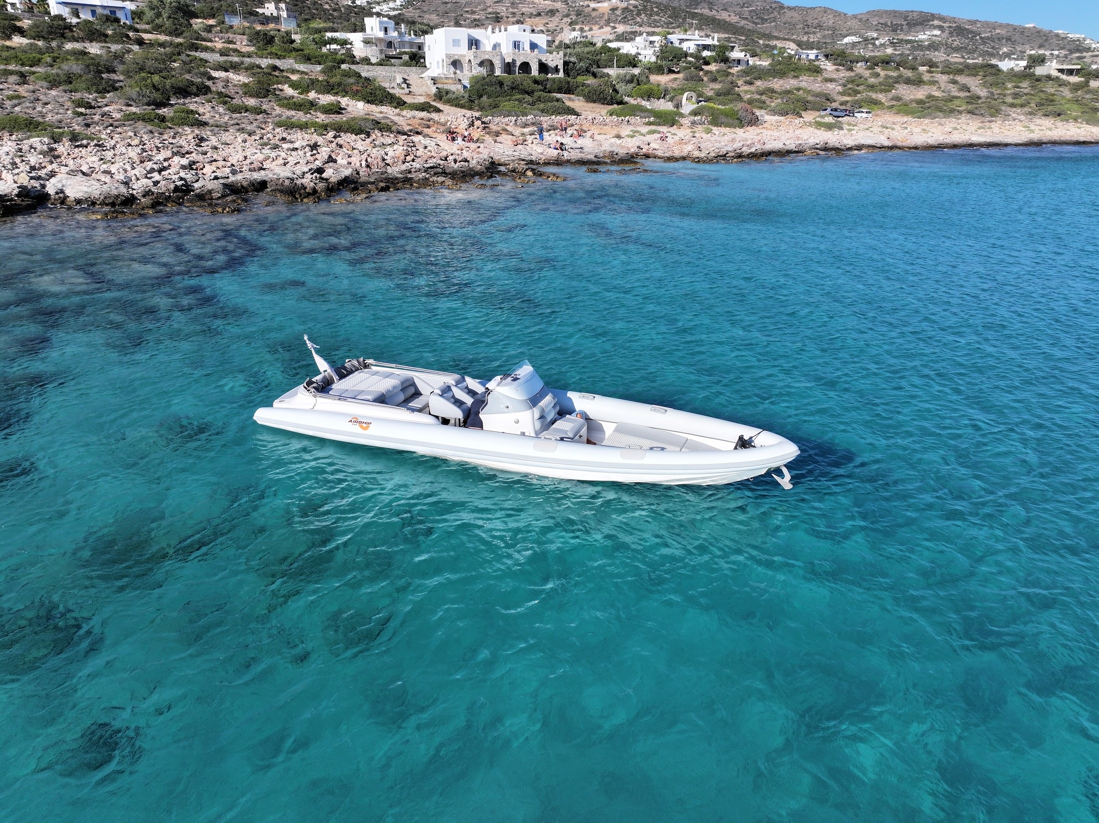 White luxury speedboat anchored in crystal-clear turquoise waters near a rocky Cycladic coastline with traditional white houses.