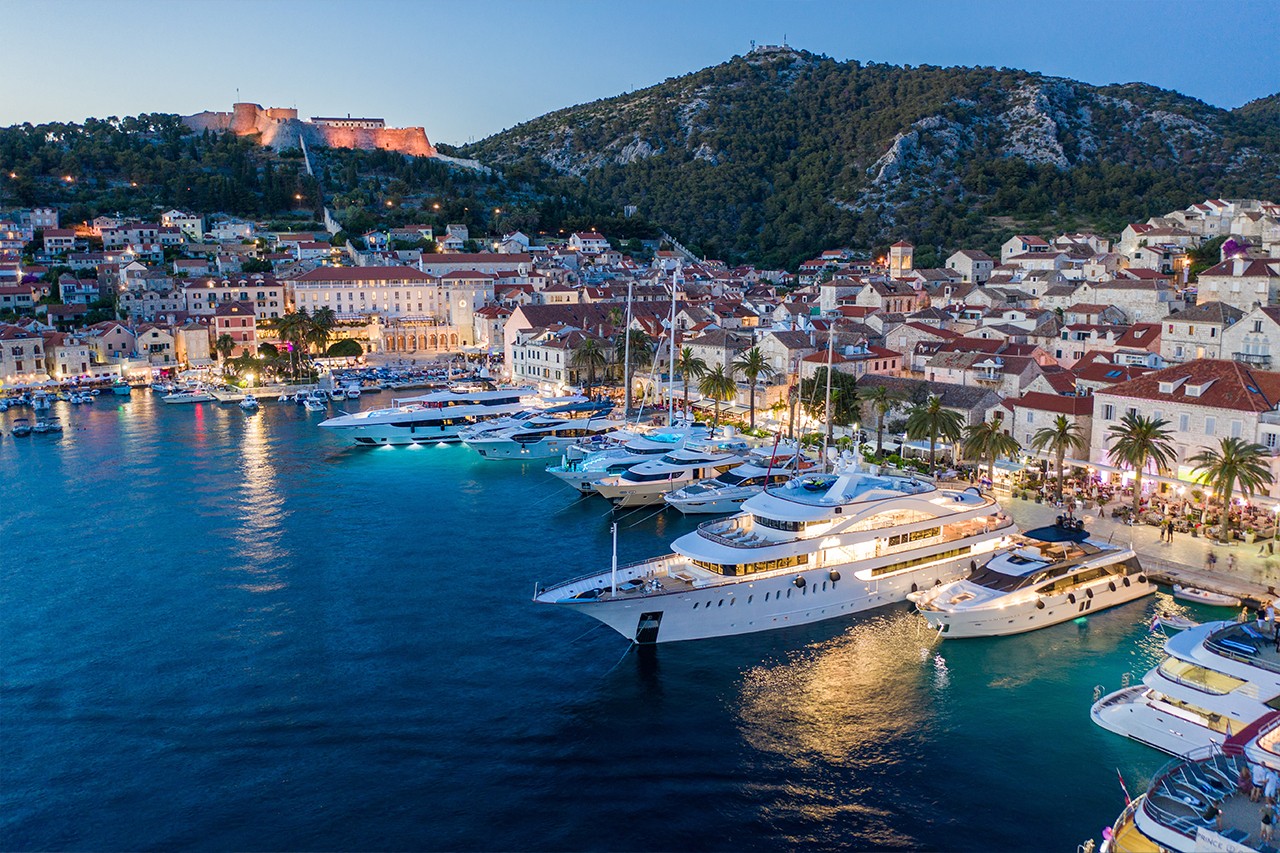 Boats parked in a Croatian marina at twilight, with a small city, fortress and hills in the background