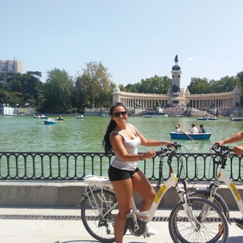 Güneşli bir günde teknelerle dolu bir gölün yakınında bisikletle duran bir kadın; arka planda anıt ve ağaçlar.