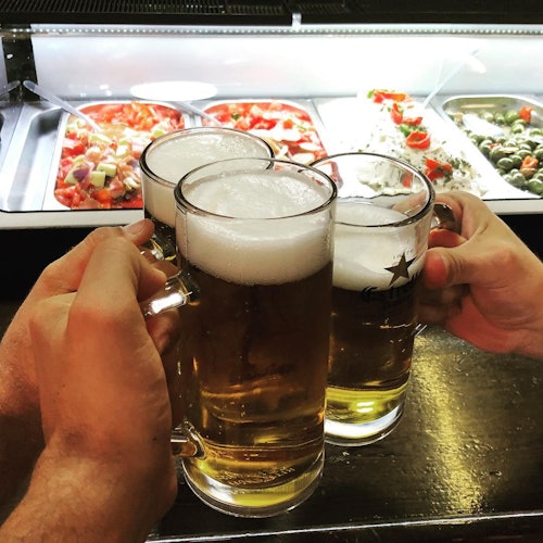 Three hands clinking mugs of beer in front of a salad bar offering various dishes.