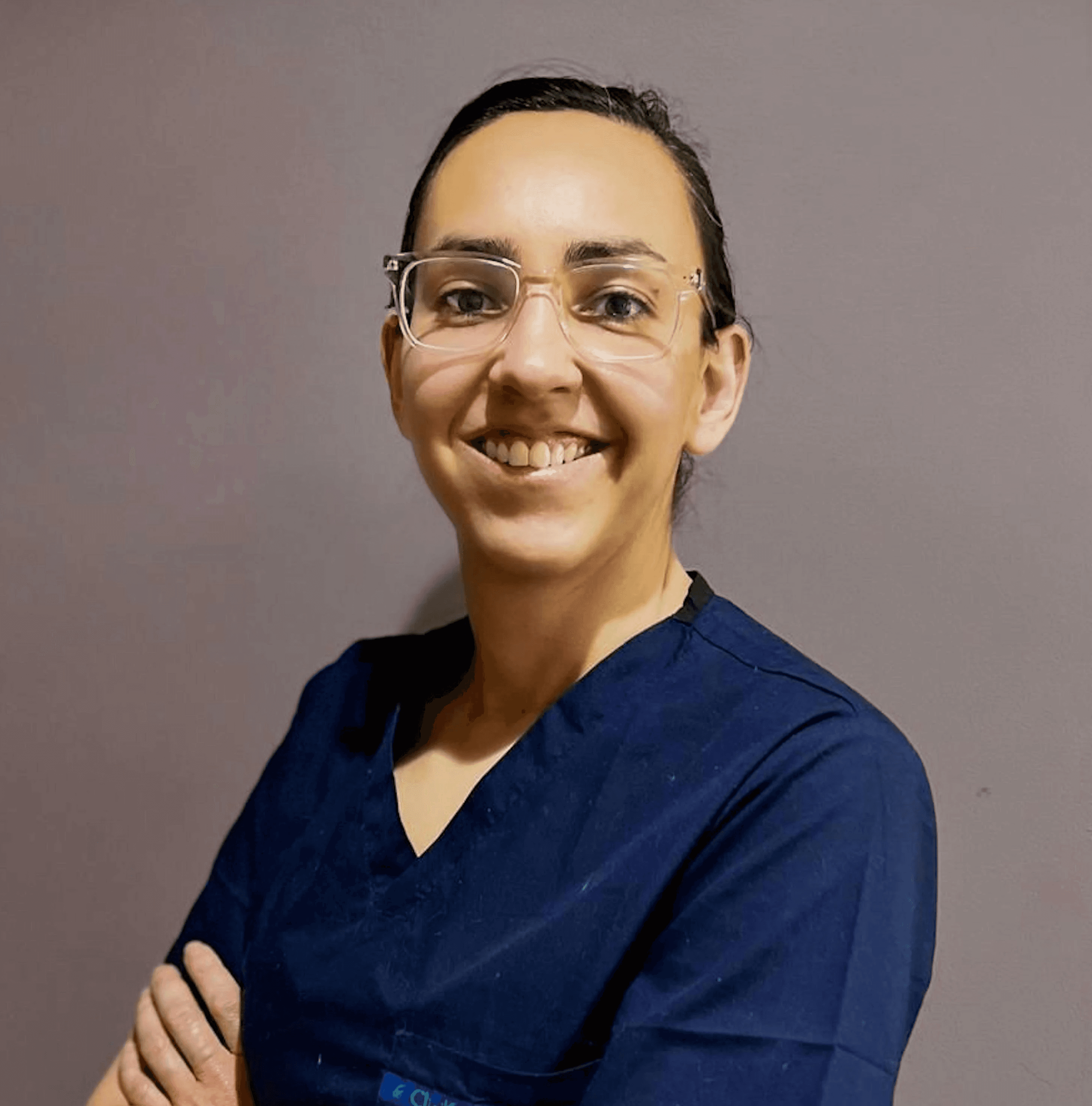 A woman in blue scrubs with glasses smiles.