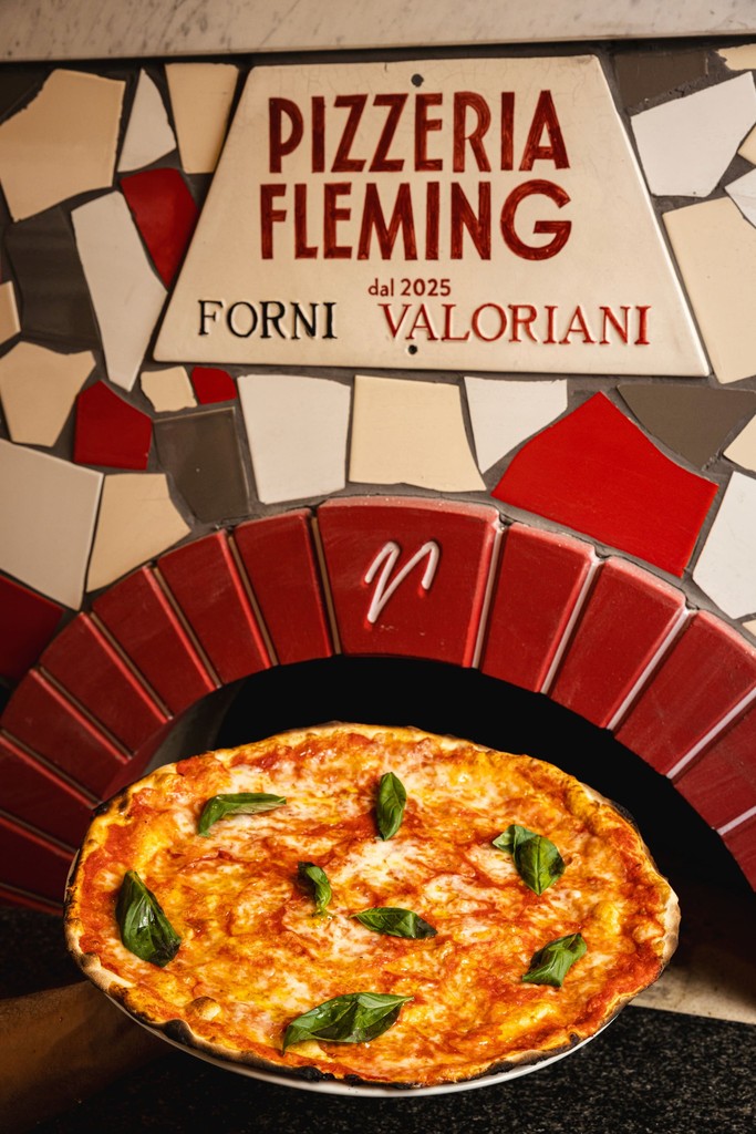 A delicious dish of seafood pasta sits in a bowl, with a brick oven and restaurant sign in the background.