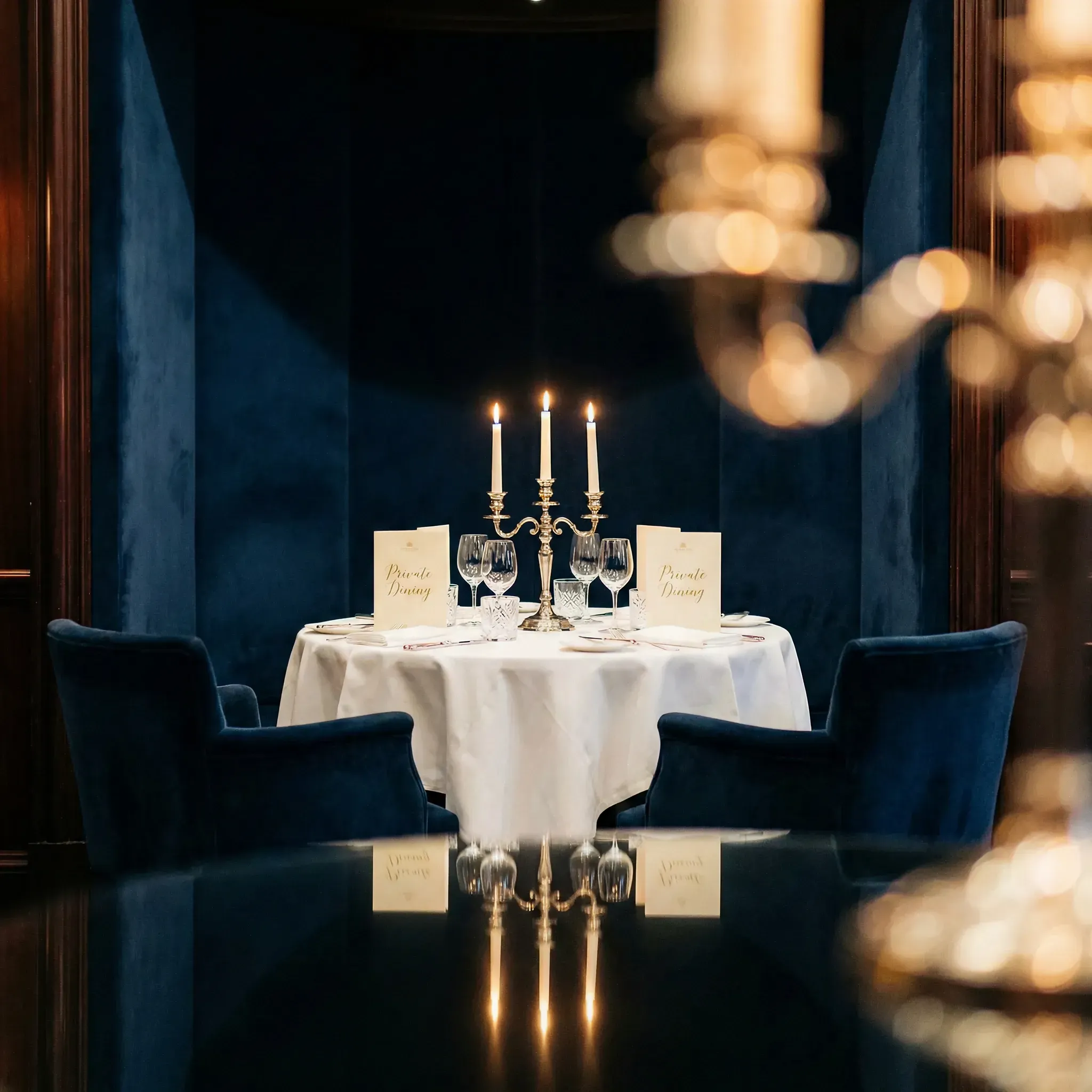 Elegant dining scene with a round table set for two, featuring a white tablecloth, three lit candles, and blue velvet chairs in a dimly-lit, plush setting.