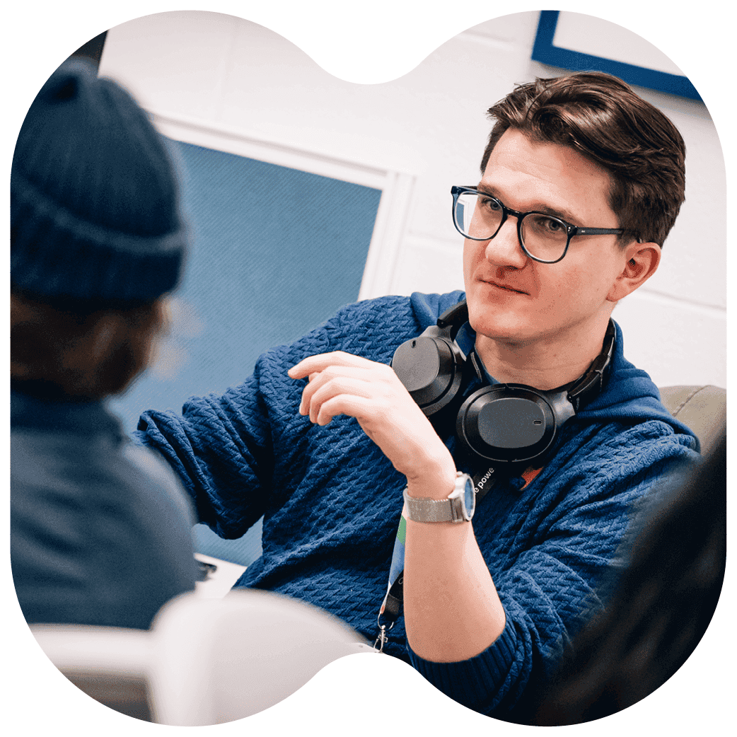 Person seated in a discussion, wearing headphones around the neck and gesturing while listening to another person.