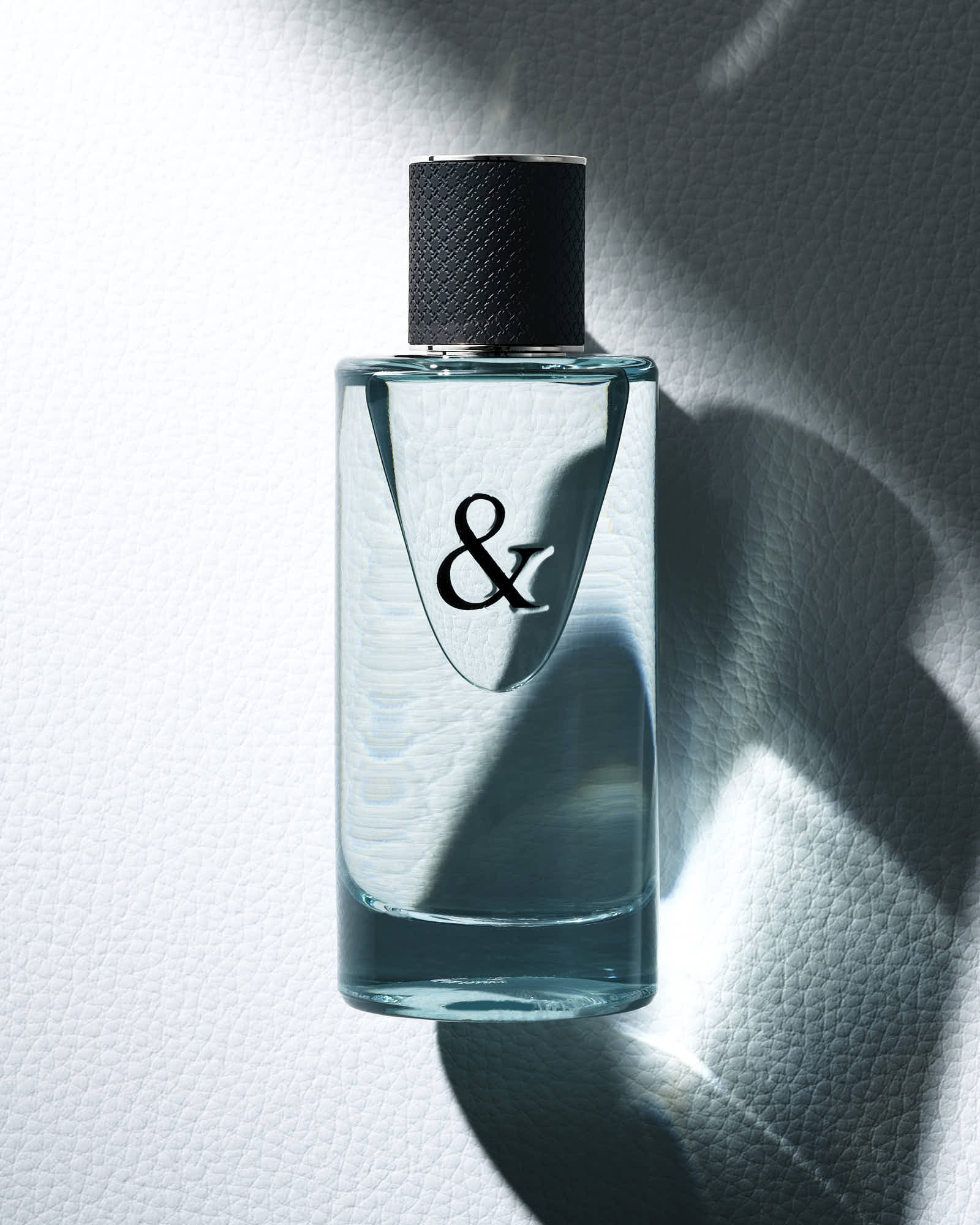 A clear perfume bottle with a black cap, featuring a minimalist design, is illuminated against a soft shadowy background.