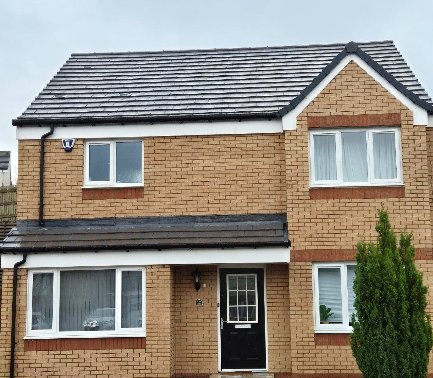A modern two-story brick house with a dark tiled roof. It features three white-framed windows and a black front door, surrounded by light greenery.
