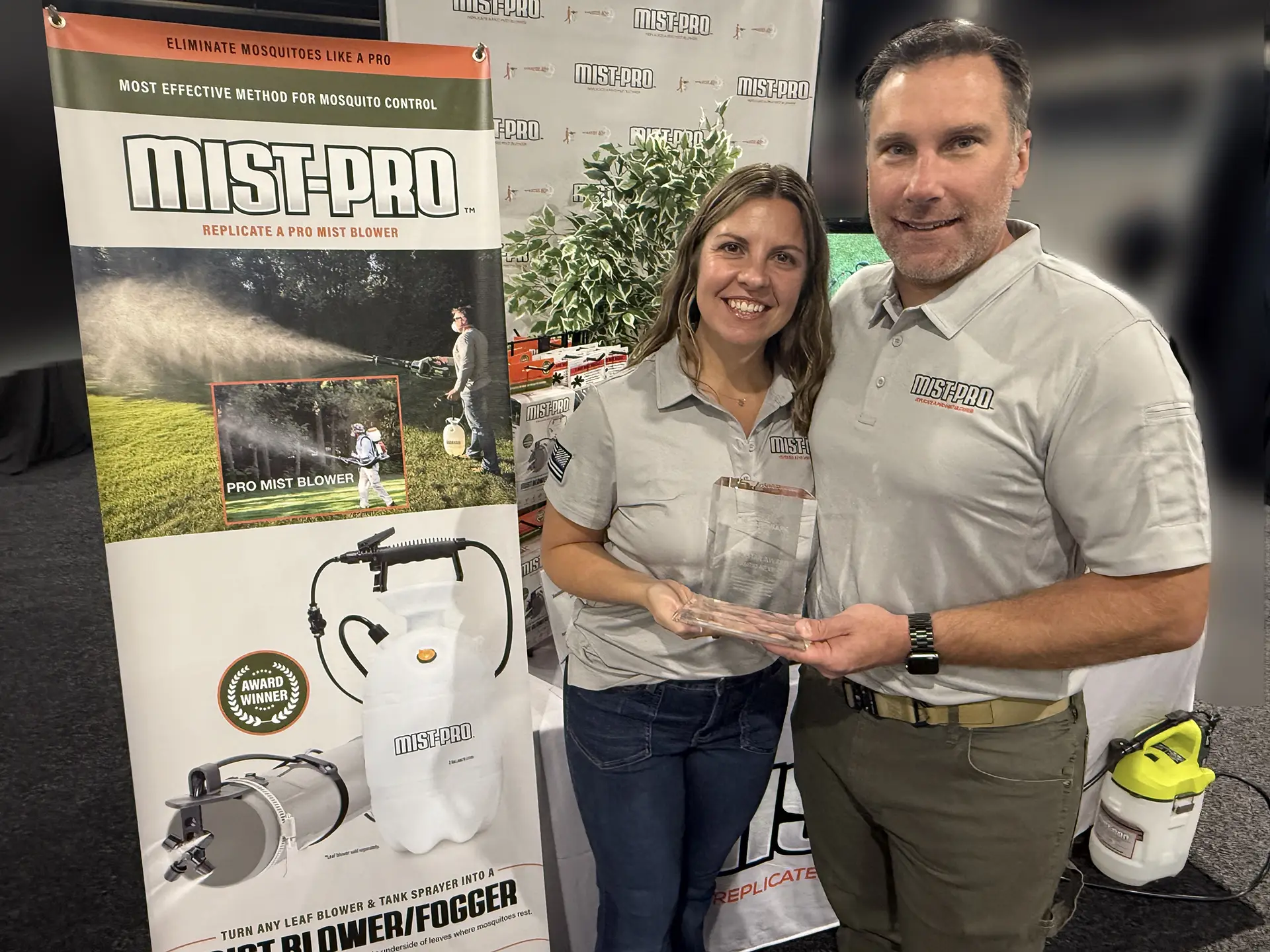 A smiling man and woman in branded gray shirts hold an award. They stand beside a "Mist-Pro" mosquito control banner at a booth. The mood is celebratory.