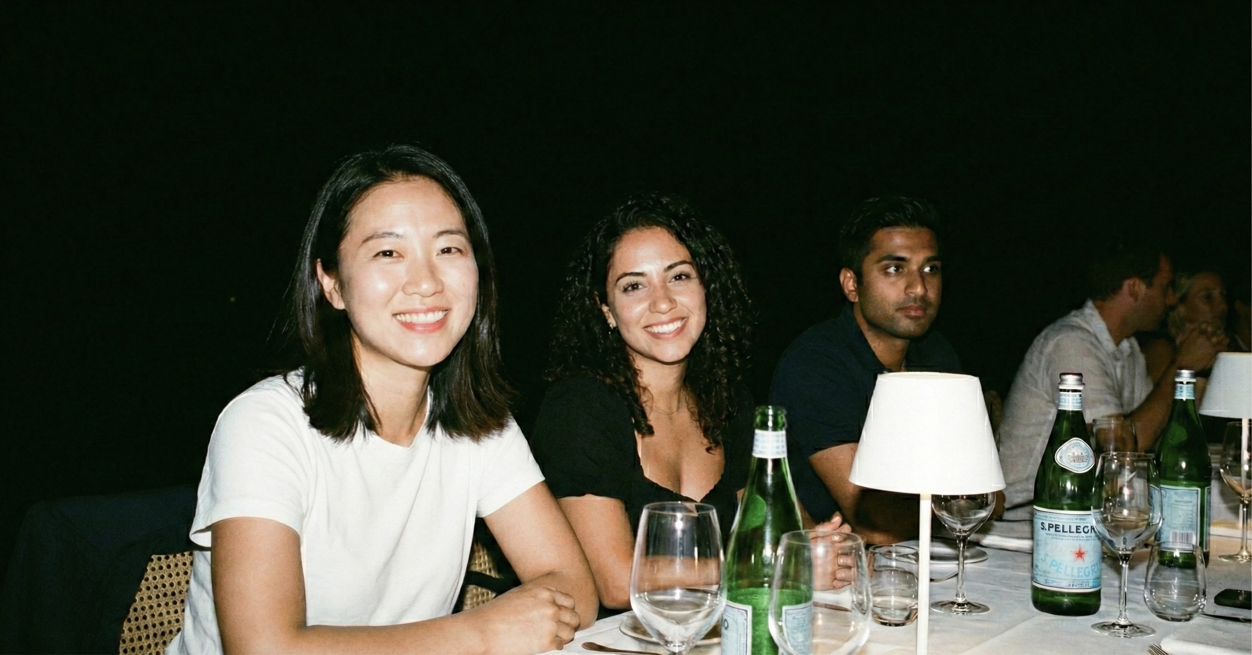 Restaurant interior with wooden tables and chairs lined against a textured wall, softly lit by round wall lamps.