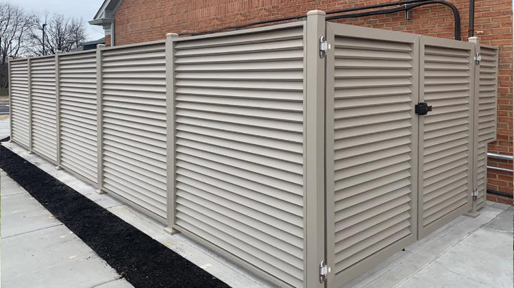 A tan louvered aluminum privacy enclosure featuring a double gate with a black latch, installed on a concrete pad next to a brick building.