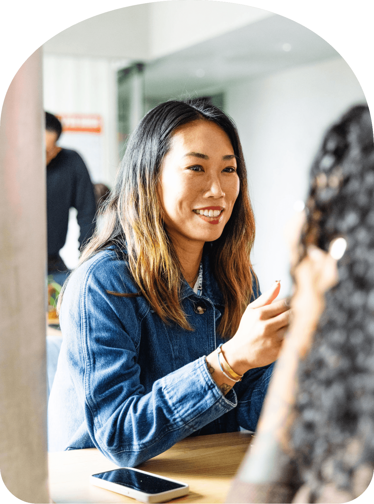 Woman smiling and gesturing while talking with a colleague at a table in a bright, casual office setting.
