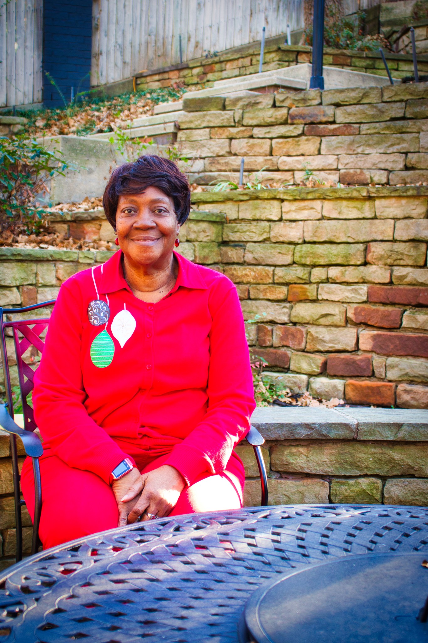 A cheerful elderly Black woman in a bright red outfit sits at a patio table, wearing festive ornament-shaped pins and smiling warmly at the camera, with a stone wall and steps in the background