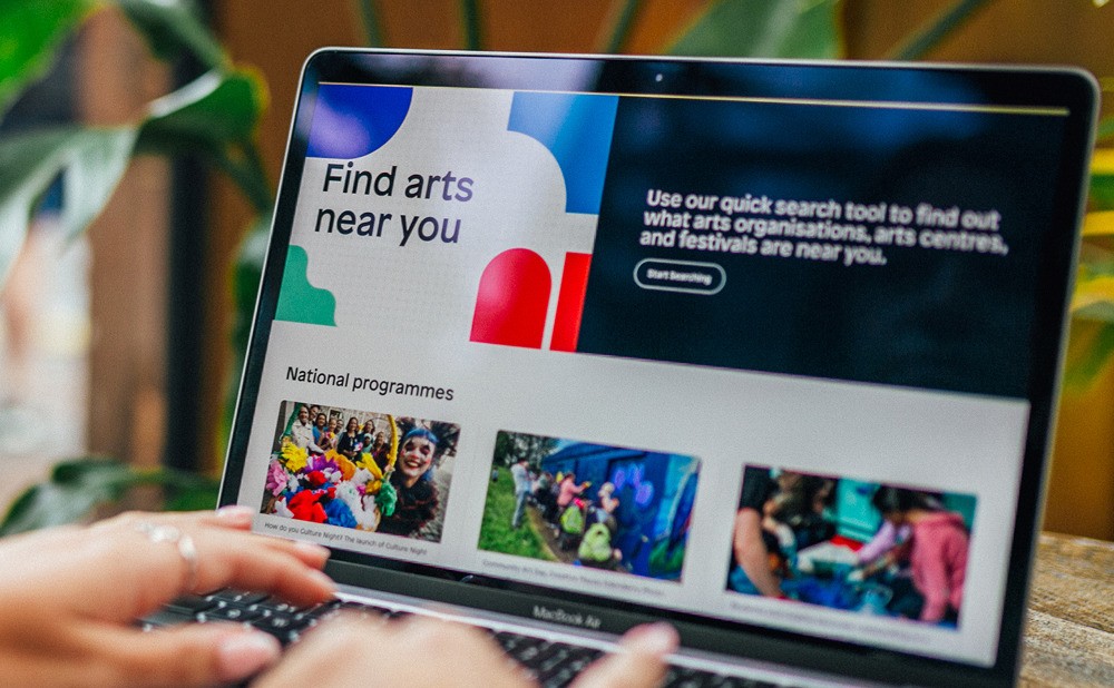 Person using a laptop displaying the Arts Council of Ireland website homepage, featuring art event listings and colourful design elements.