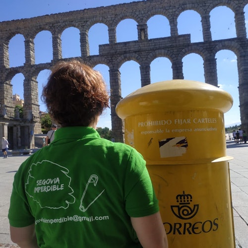 A person in a green shirt stands next to a yellow mail bin in front of an ancient stone aqueduct.