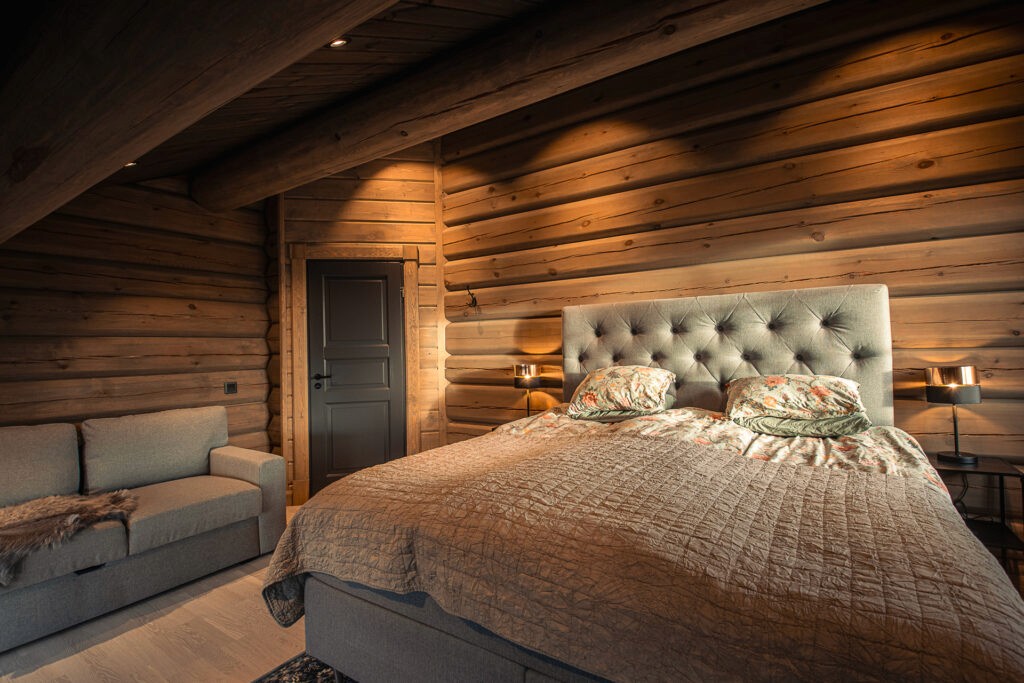 A rustic log house bedroom with a sloped ceiling, a double bed, and exposed round timber ceiling beams, creating a warm and inviting atmosphere.