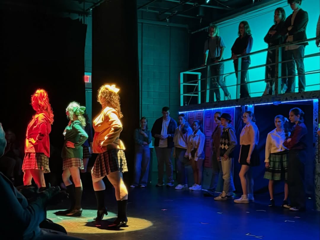 The three Heathers stand downstage in individual top spotlights, each a different color — red, green, and yellow — to match the signature colors of each Heather. Upstage, in front of and on a platform, the remaining cast stands looking out at them. The cyc behind the platform is turquoise, and the stage is washed in a subtle blue.