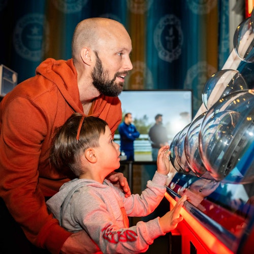 En skægget mand og et barn interagerer med en videnskabsudstilling, oplyst af rødt lys, med et gruppefotografi i baggrunden.