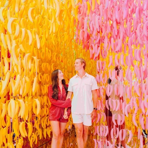 一对情侣在一个黄色和粉色的香蕉主题房间里，手牵手微笑着互相凝视。