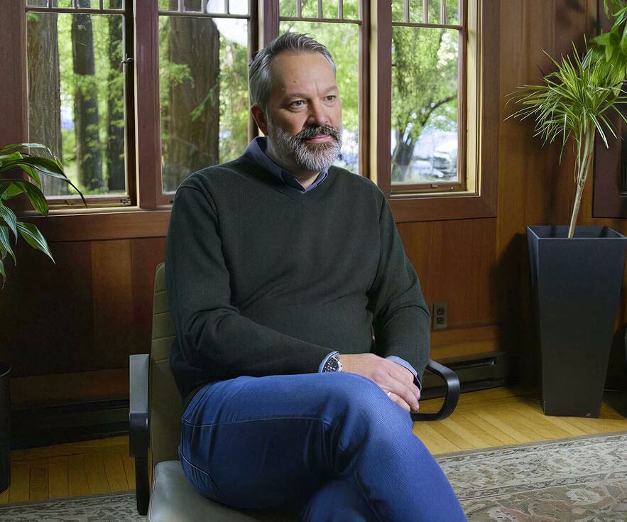 Man sitting in a chair with a window and trees in the background looking relaxed.