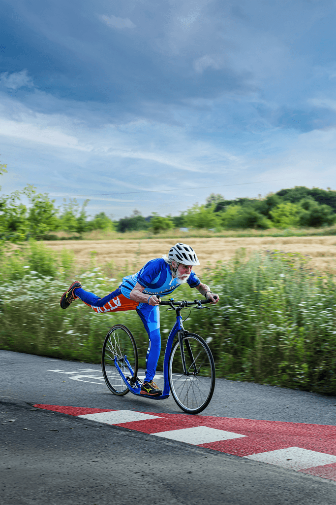 Mężczyzna na niebieskim rowerze-hulajnodze z podniesioną nogą, w kasku, na tle łąki i nieba. Sport i aktywność na świeżym powietrzu.