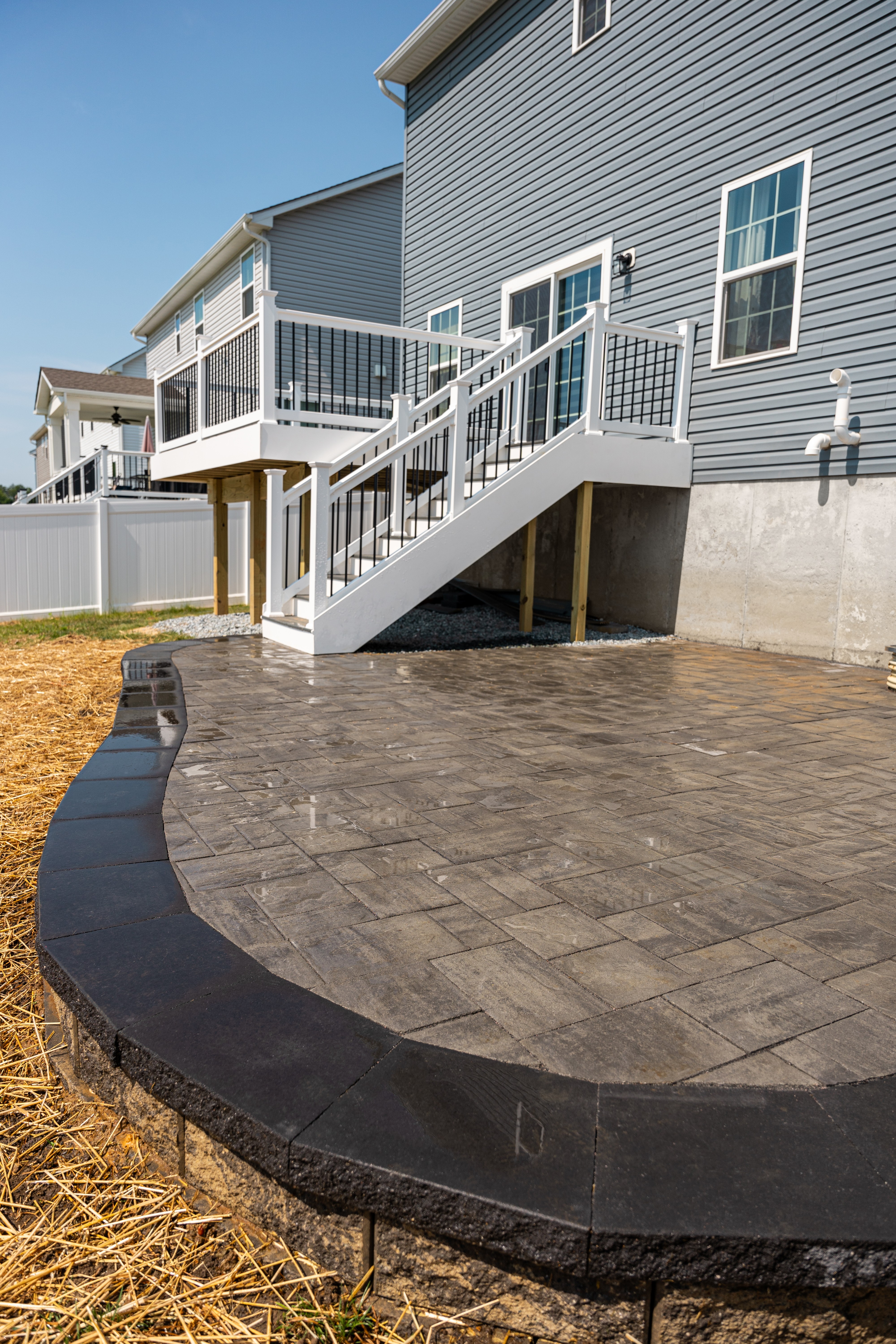 A newly constructed multi-level deck with white railings leads down to a spacious grey stone patio area in a backyard, surrounded by manicured soil and straw, against the backdrop of a blue, two-story house.
