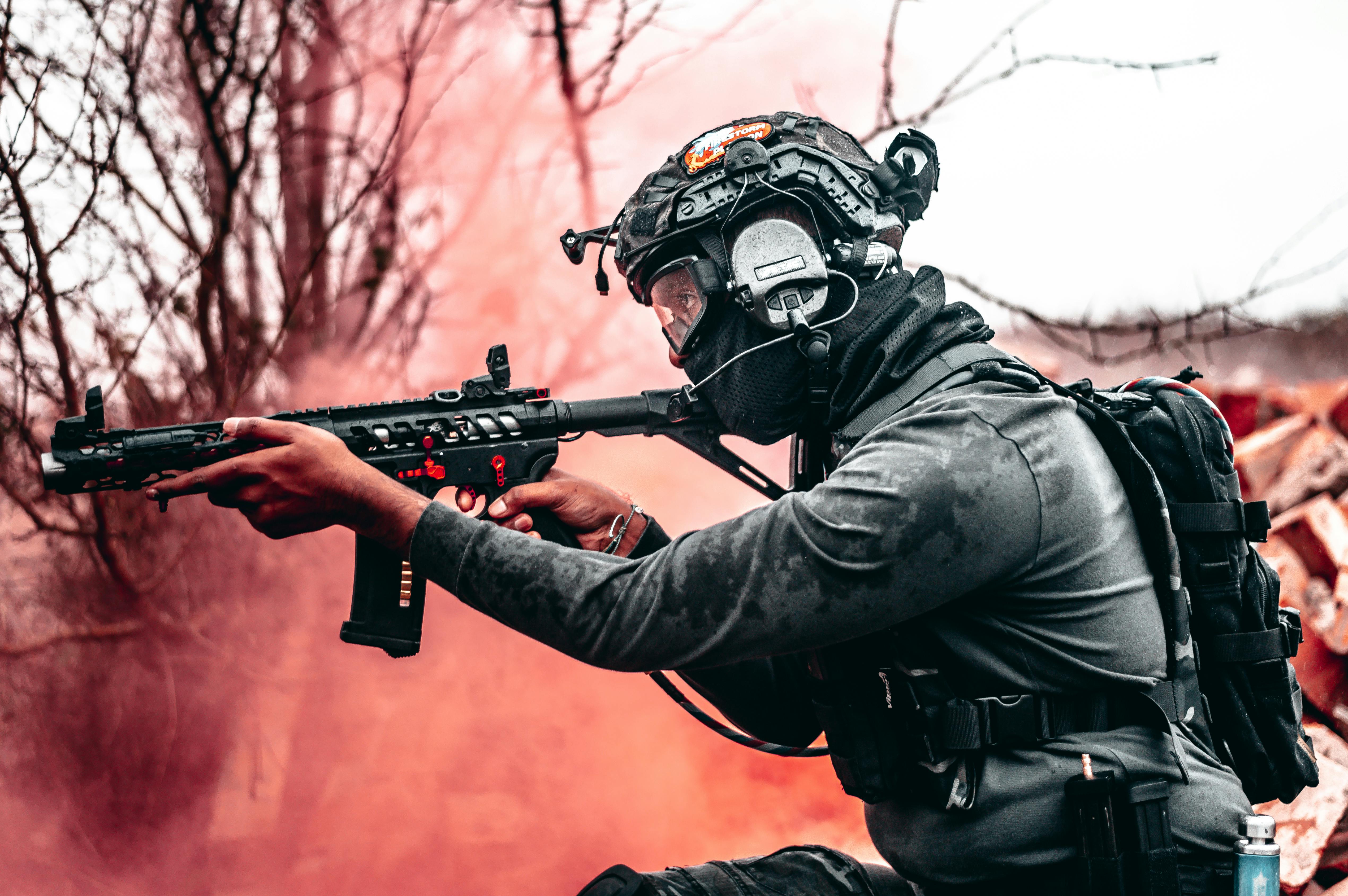 A tactical operator in a kneeling position holding an assault rifle. The person is wearing a helmet with mounted night vision equipment, tactical gear, and dark clothing. The image shows someone in full combat equipment posed against a blurred background.