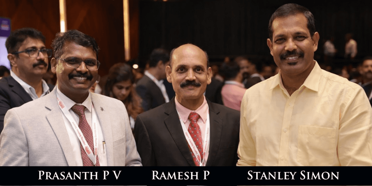 Three men in business attire stand smiling at an event. They are in a crowded room with blurred figures in the background, conveying a professional and friendly atmosphere.