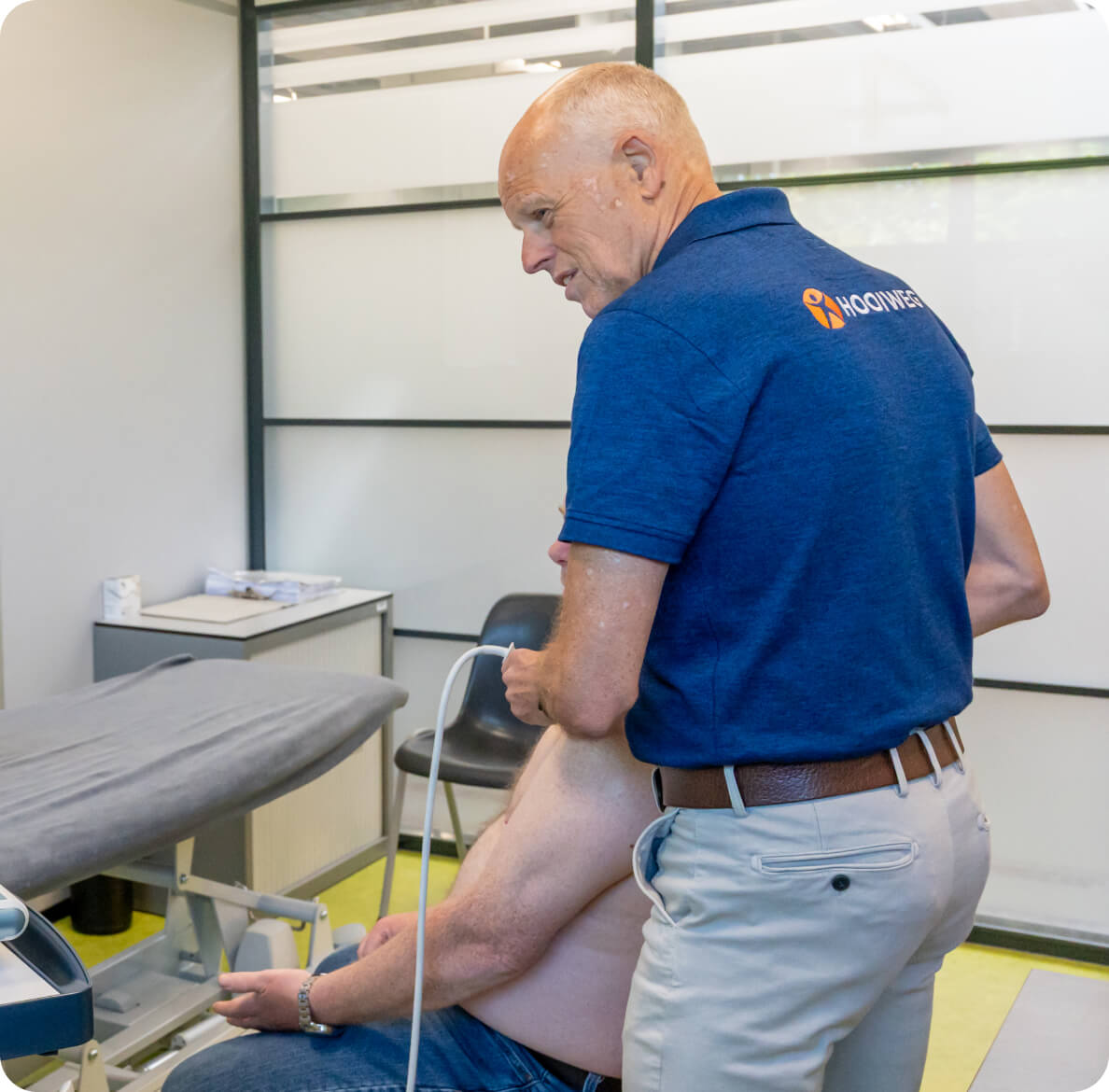 A chiropractor in a blue polo shirt performs an adjustment on a seated patient in a modern clinic, with a massage table and tidy workspace in the background.