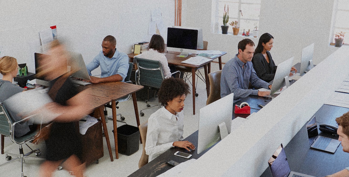 A grainy and motion blurred image of people working in an office at their computers with people walking around.