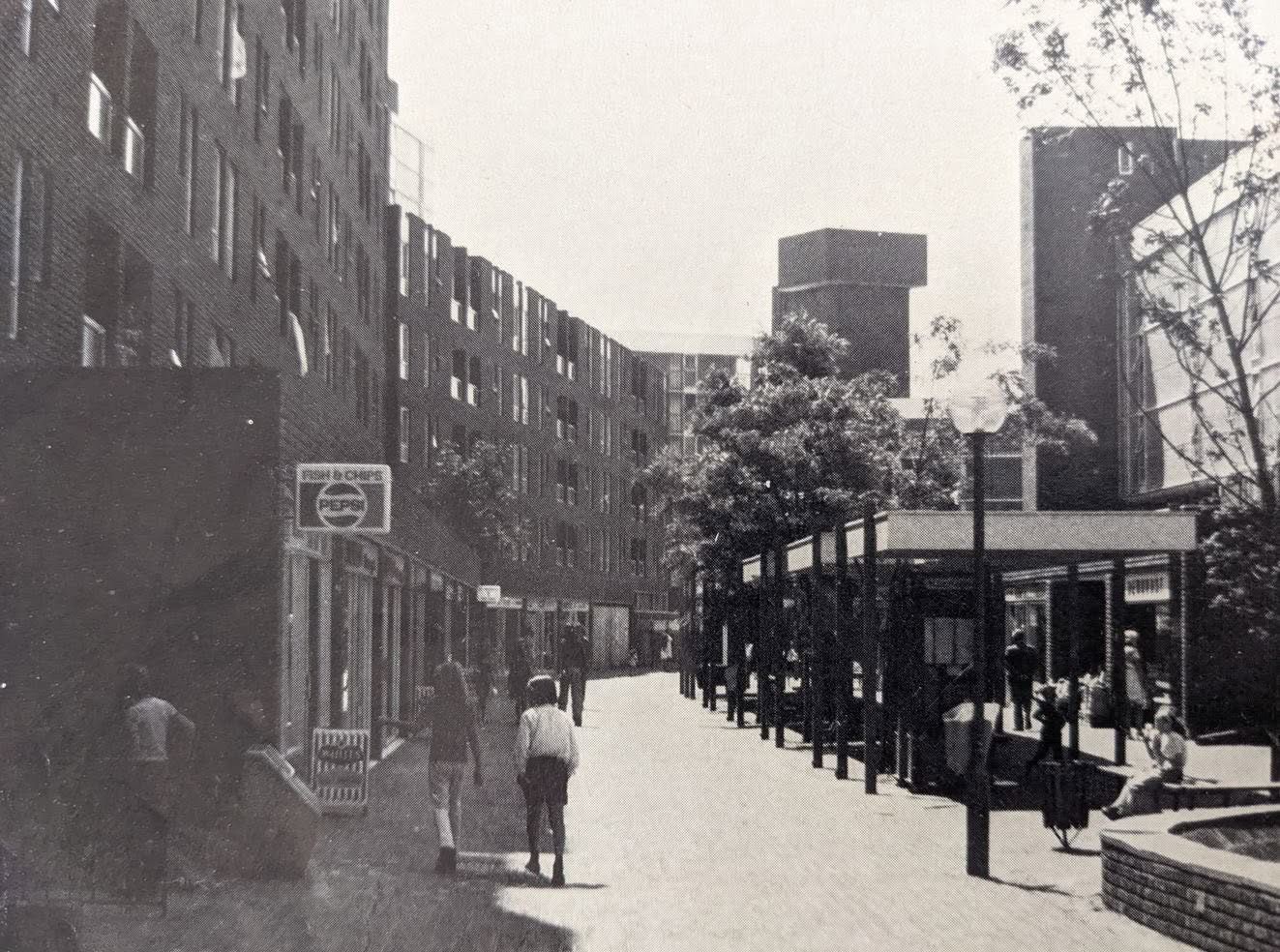 Black-and-white photograph of a pedestrian street lined with modern brick buildings and shopfronts. Several people walk along the wide paved walkway, with trees, benches, and a low pavilion-style structure on the right. A sign reading “Fish & Chips” is visible on the left, and taller buildings rise in the background.