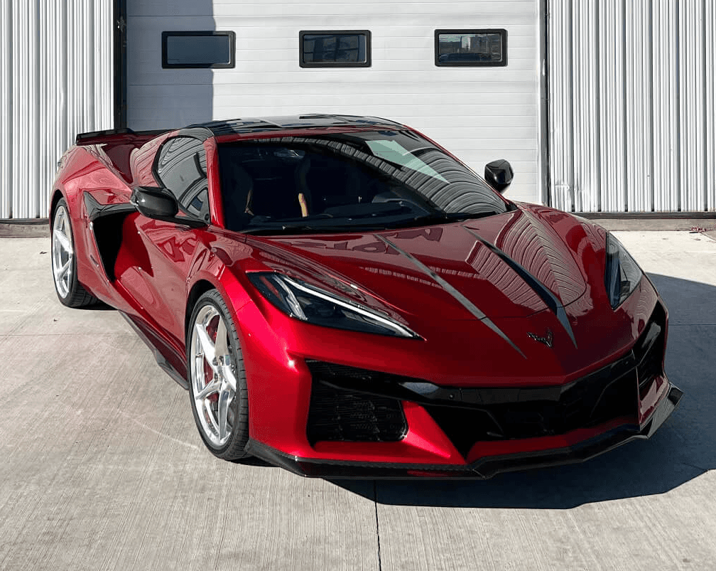 A shiny red Chevrolet Corvette sports car is parked on a concrete driveway in front of a white industrial building.