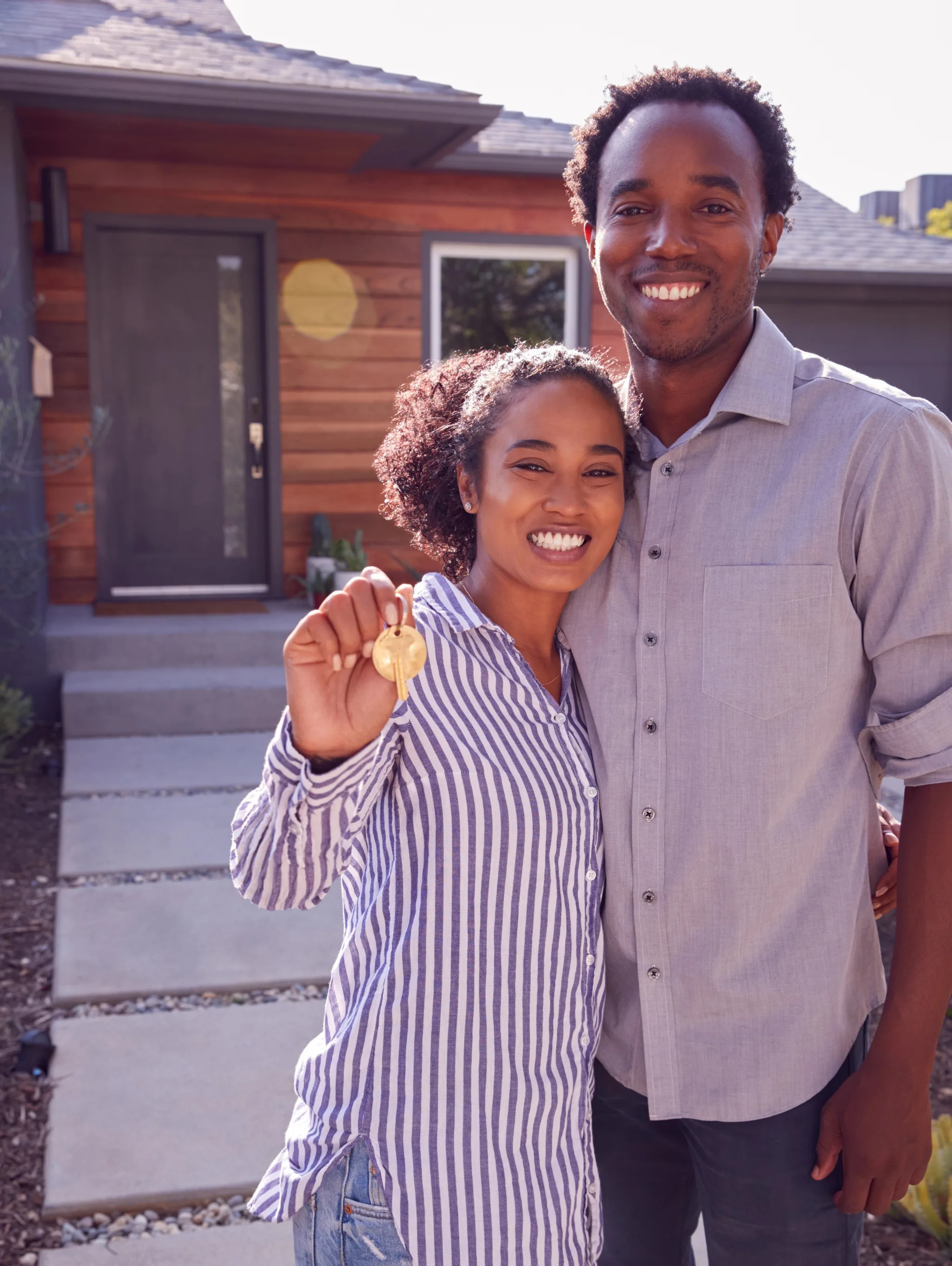 A smiling couple stands in front of their modern wooden house, with the woman holding up a gold house key to celebrate their new home purchase.