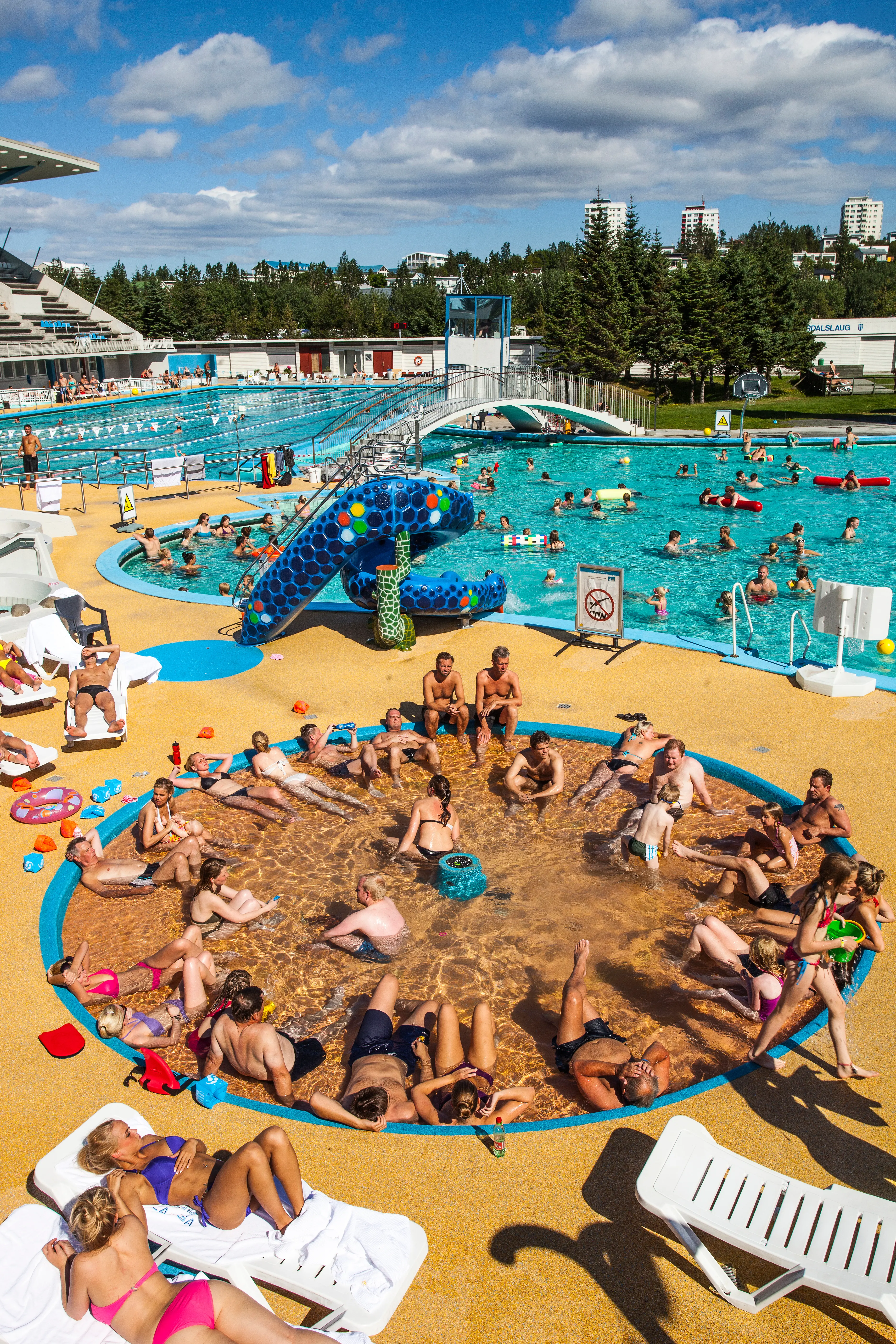 Swimmers and sunbathers enjoying the large outdoor geothermal pool at Laugardalslaug in Reykjavík on a bright summer day, with lap lanes, hot tubs, and families relaxing around the pool deck.