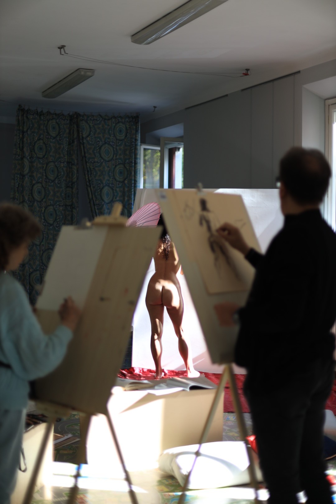 In a dimly lit art studio, artists focus on sketching a nude figure standing in a sunlit pose, with easels and canvases in the foreground.