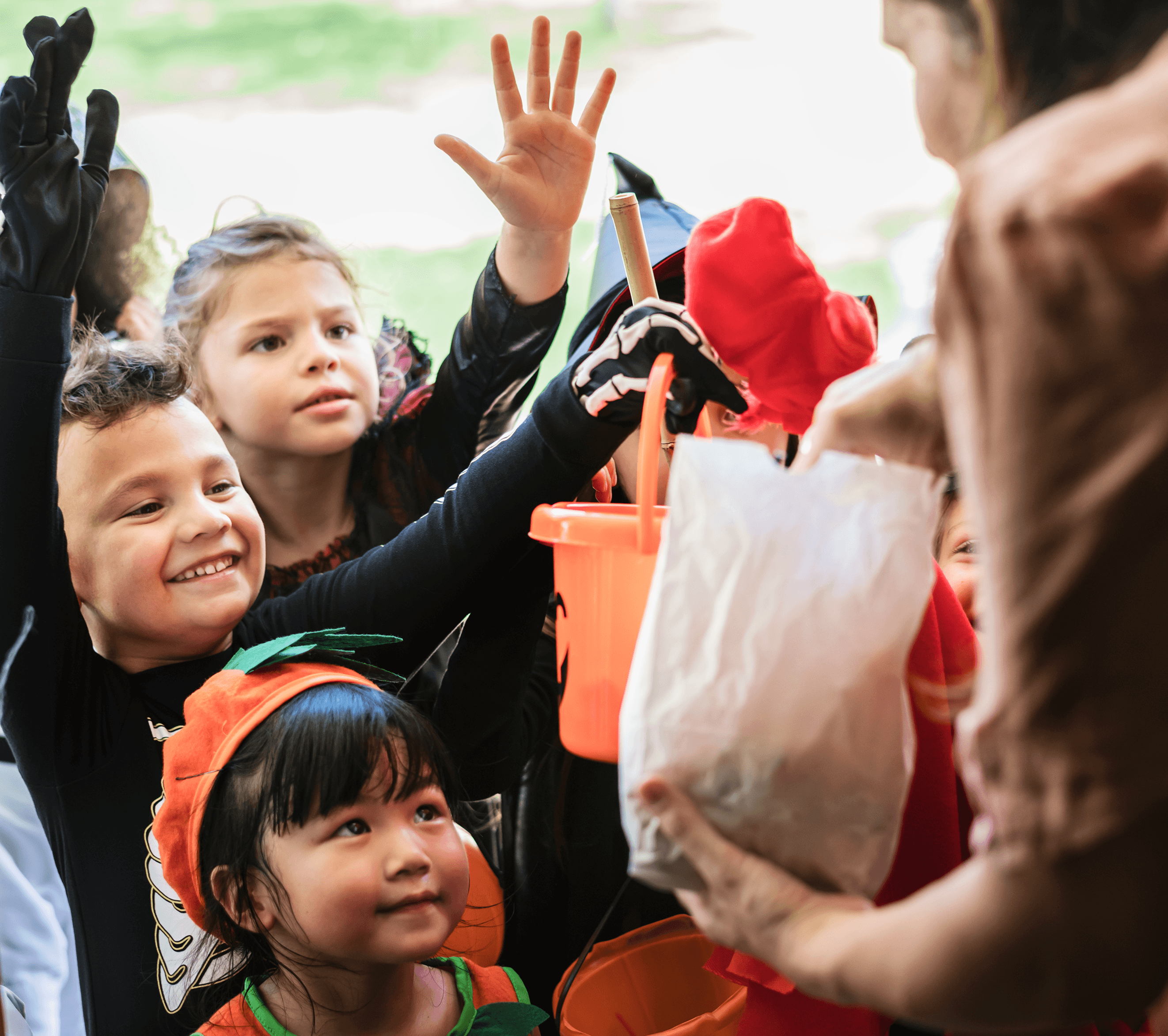 Children eagerly reach out with hands raised as an adult hands out treats. Bright, festive decorations visible.