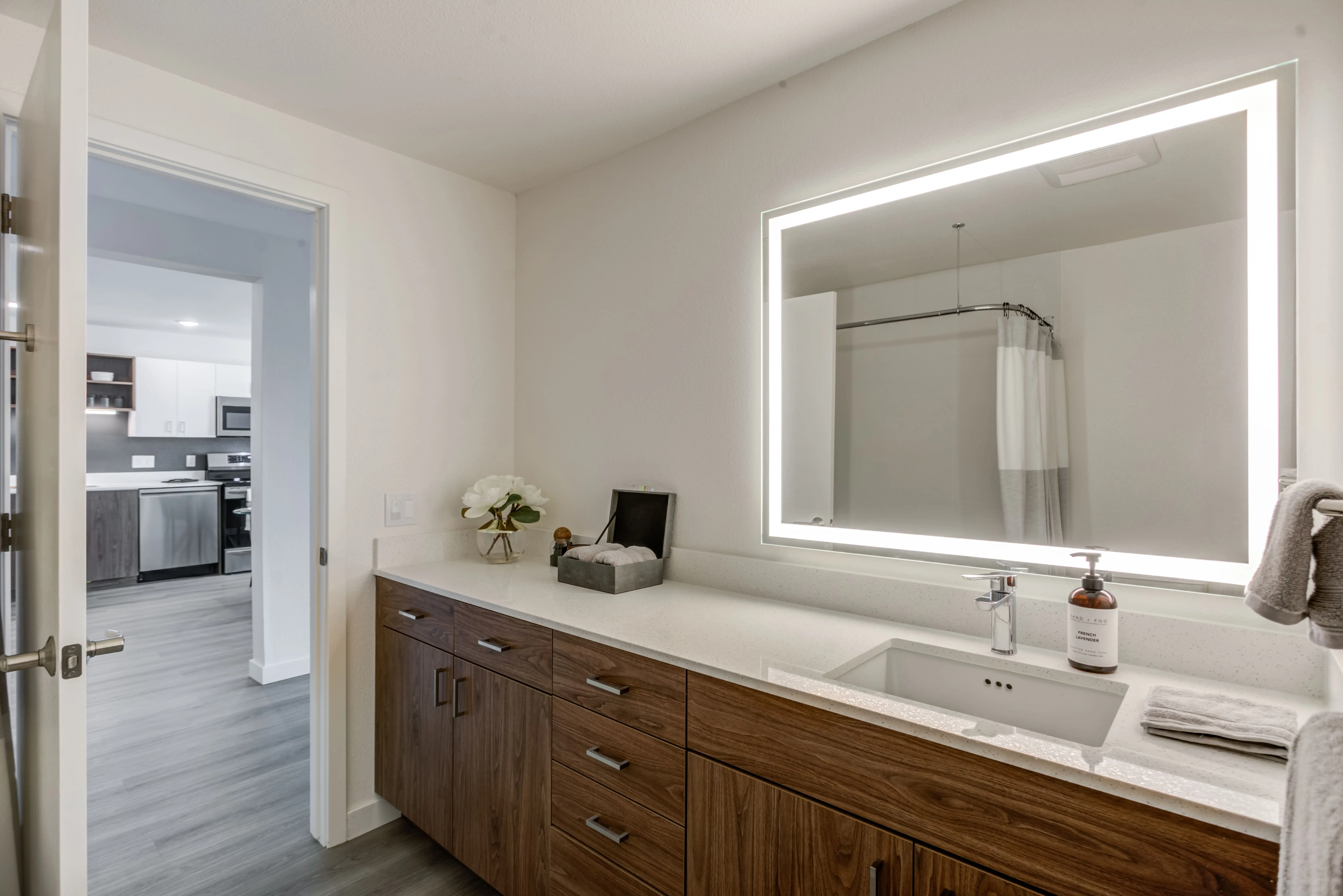 Modern bathroom featuring a large mirror with built-in lighting above a wooden vanity with multiple drawers. The sun-drenched sink area includes a soap dispenser and a folded towel. The doorway leads to a view of the kitchen in Midtown Sacramento, complete with stainless steel appliances and white cabinetry.