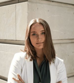 A young woman with long brown hair, wearing a white blazer and dark green shirt, stands confidently against a stone wall, creating a modern and professional atmosphere.