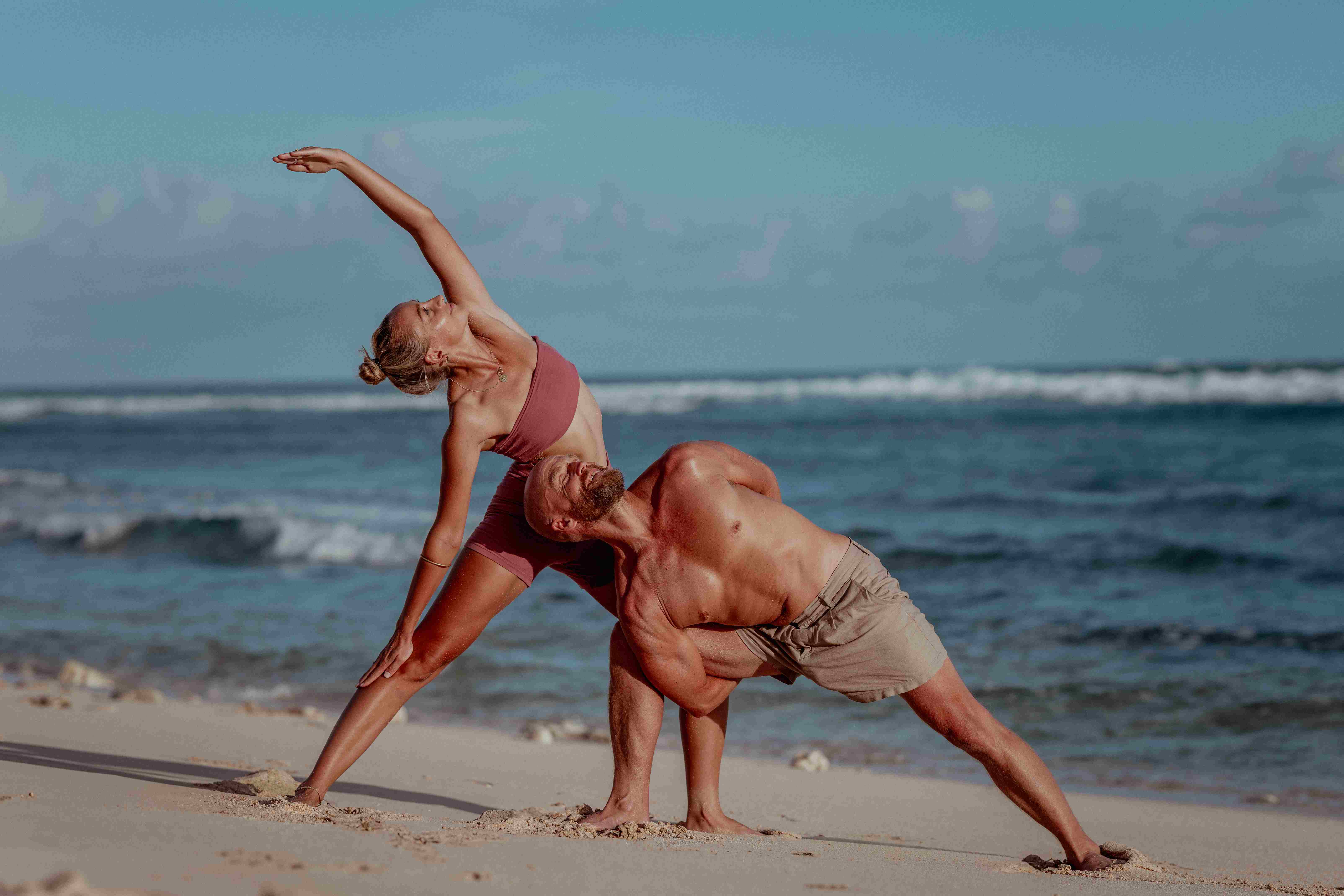 Couple practicing partner yoga side stretch on the white sand beach in Uluwatu Bali.