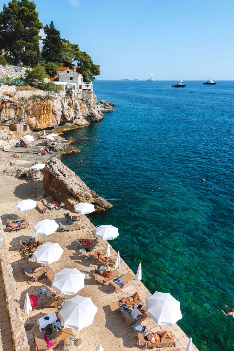 A rocky coastline with turquoise water, white beach umbrellas, and a white house on a cliff under a clear blue sky.