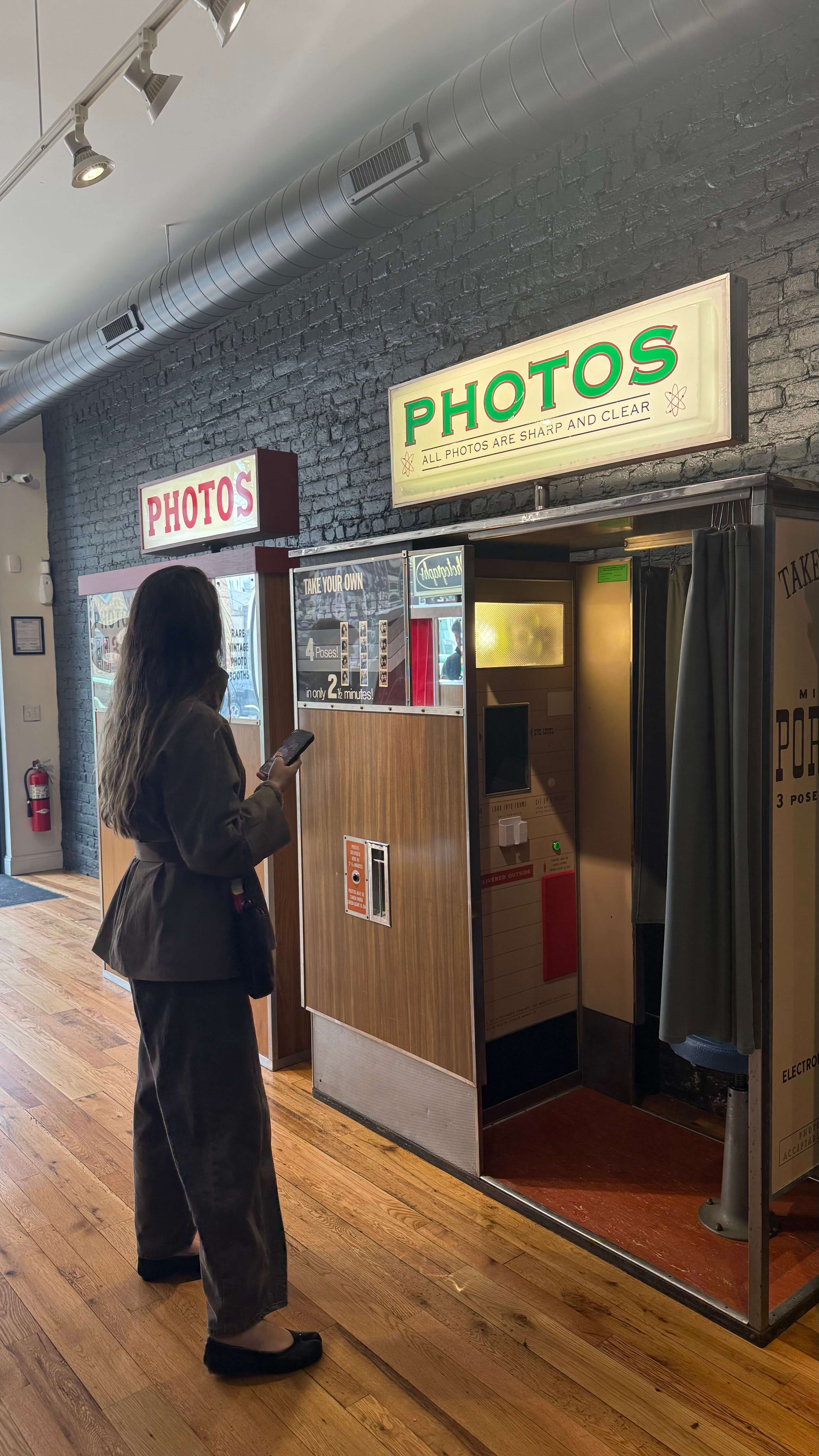 a woman with long dark hair wearing. a black pant suit stands in front of a vintage photo booth