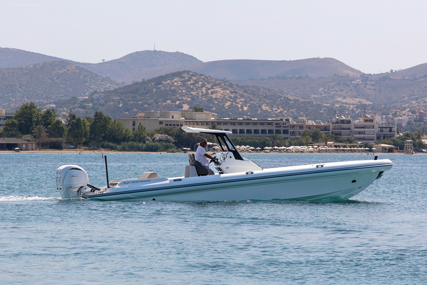 White rigid inflatable boat with twin outboard engines cruising calm blue waters with Greek coastal mountains in background.