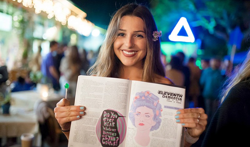 A photo of a women standing in the street at night with a lollipop and an opened book in her hands. There is a drawing of a woman in the book and is surrounded by text.