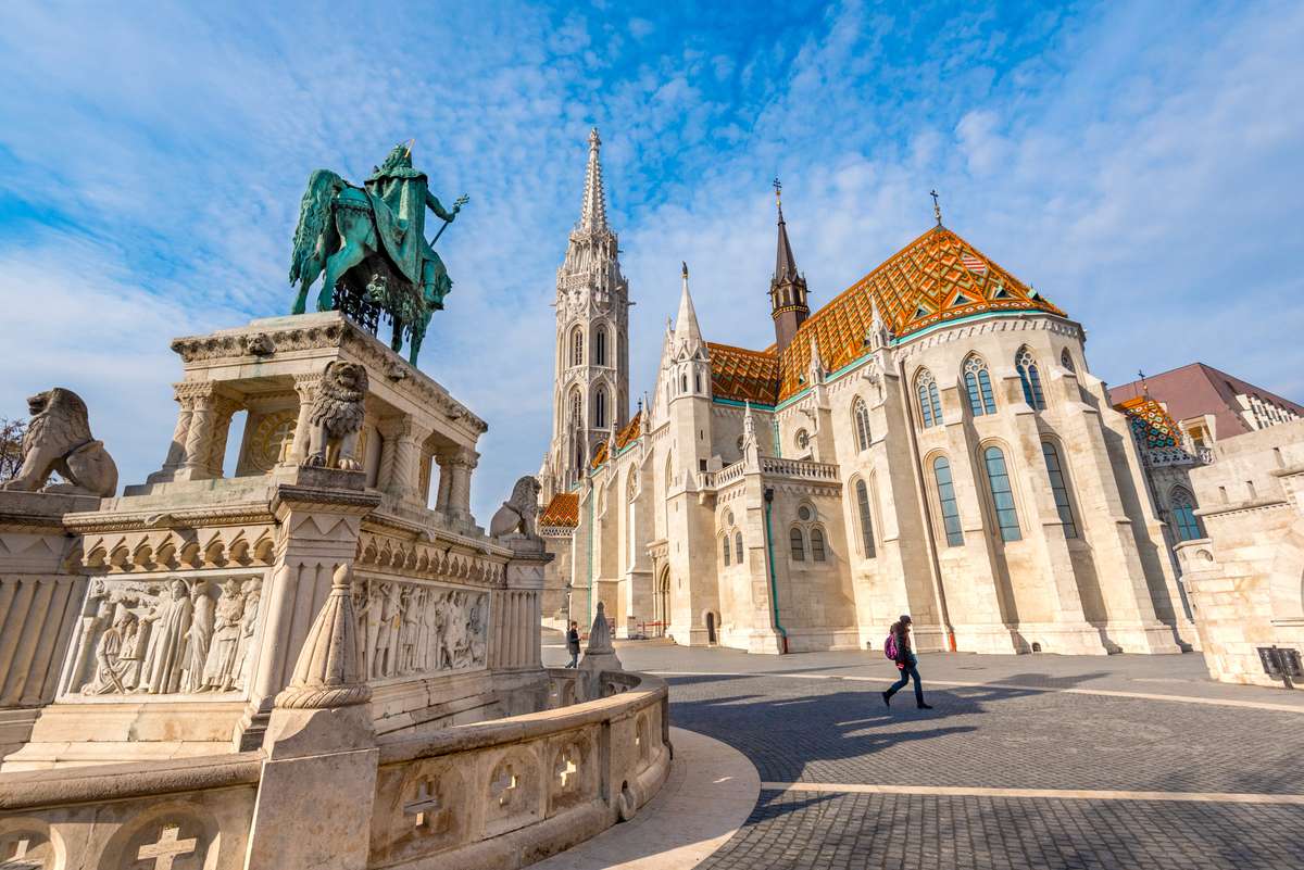 Grand architectural structures line a wide stone plaza under bright daylight, featuring vibrant blue sky and detailed stonework.