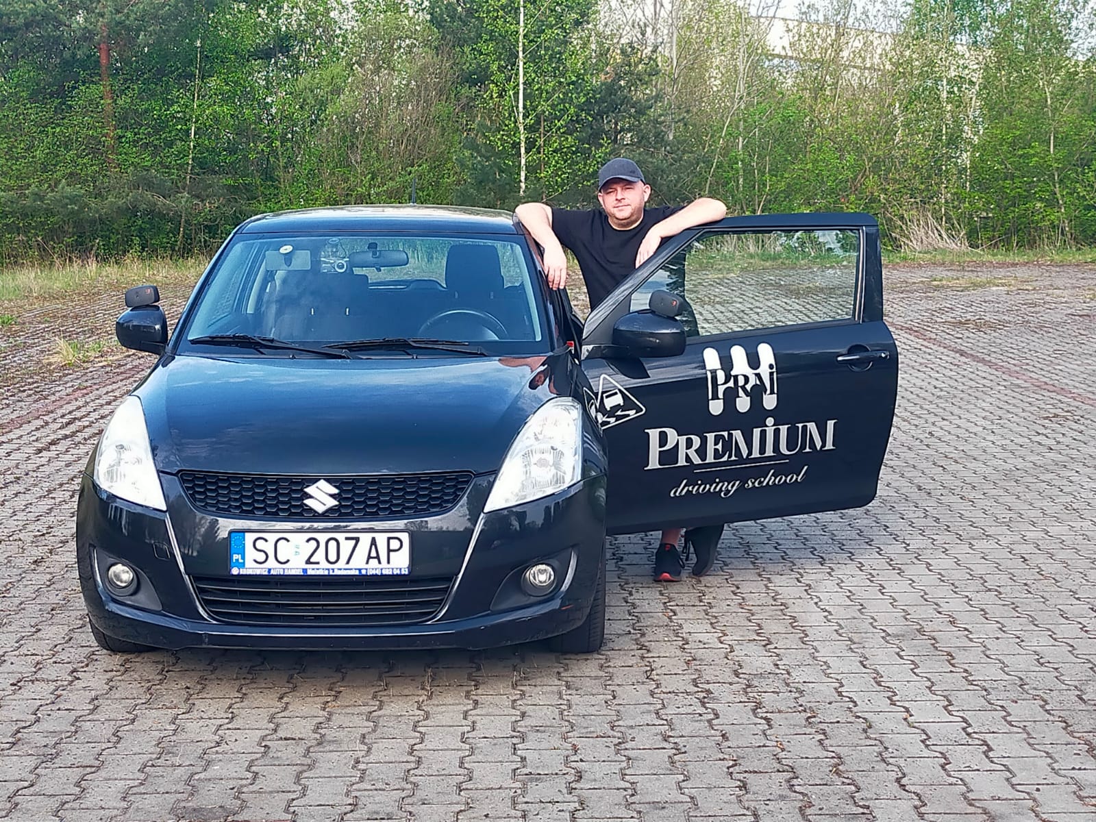Piotr Michoń stoi przy czarnym Suzuki z logo Premium, z otwartymi drzwiami.